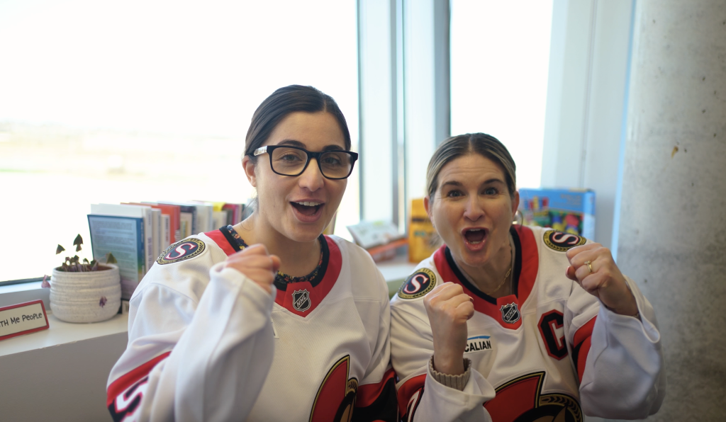 Two women wearing matching hockey jerseys cheer with raised fists while standing indoors near a window and a shelf of books.